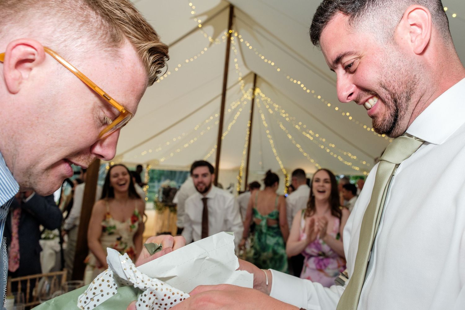 Two grooms laughing and exchanging rings at a wedding reception under a marquee tent with guests celebrating behind them.