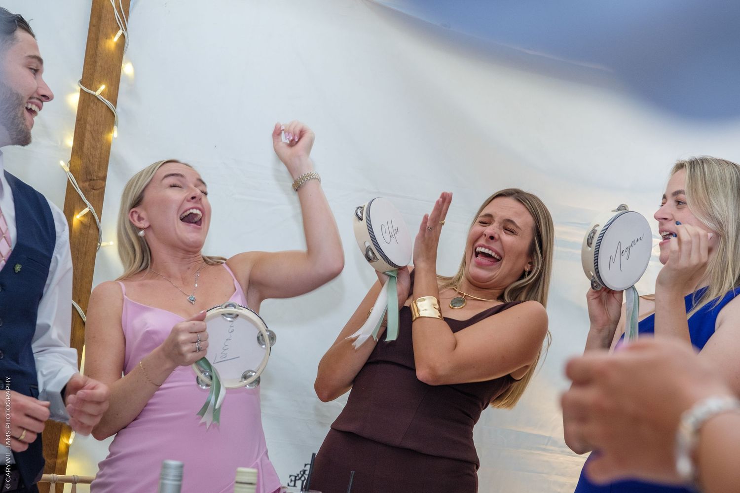 Women laughing and playing tambourines at a lively wedding reception celebration inside a tent.