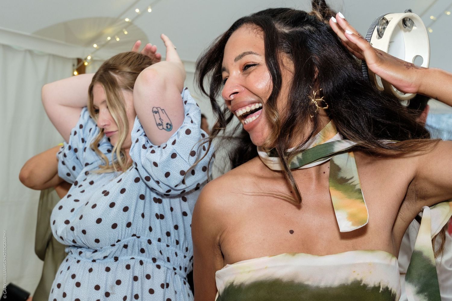 Two women dancing and laughing joyfully at a party, one in a blue polka dot dress, the other in a tie-dye top.