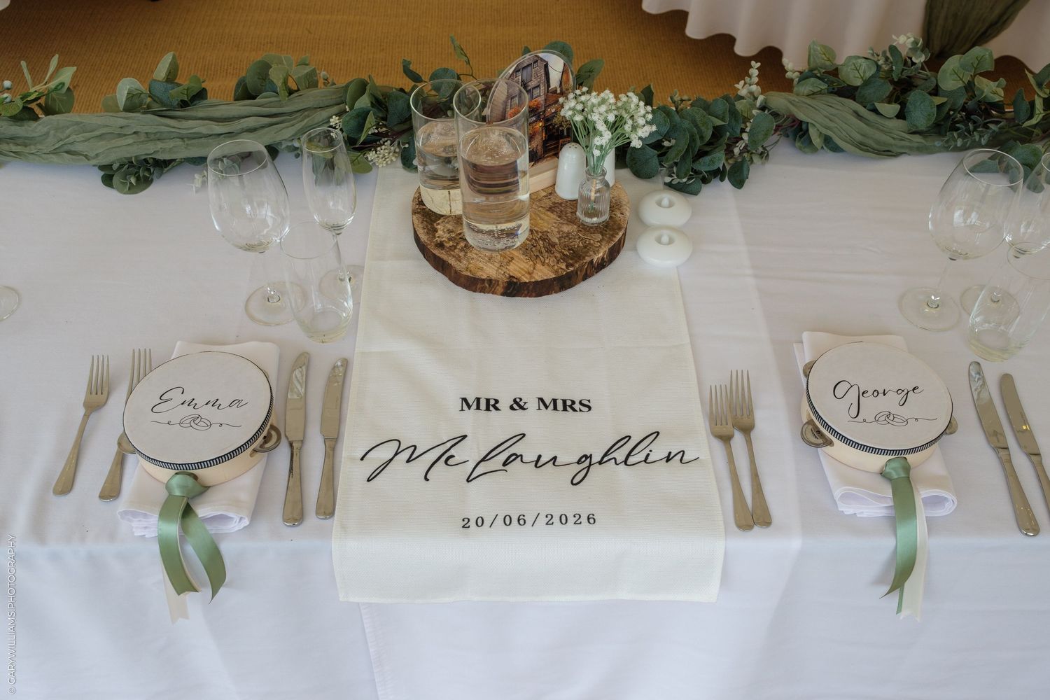 Elegant wedding table setting with Mr & Mrs McLoughlin personalized runner, wooden place cards, greenery, and gold cutlery.