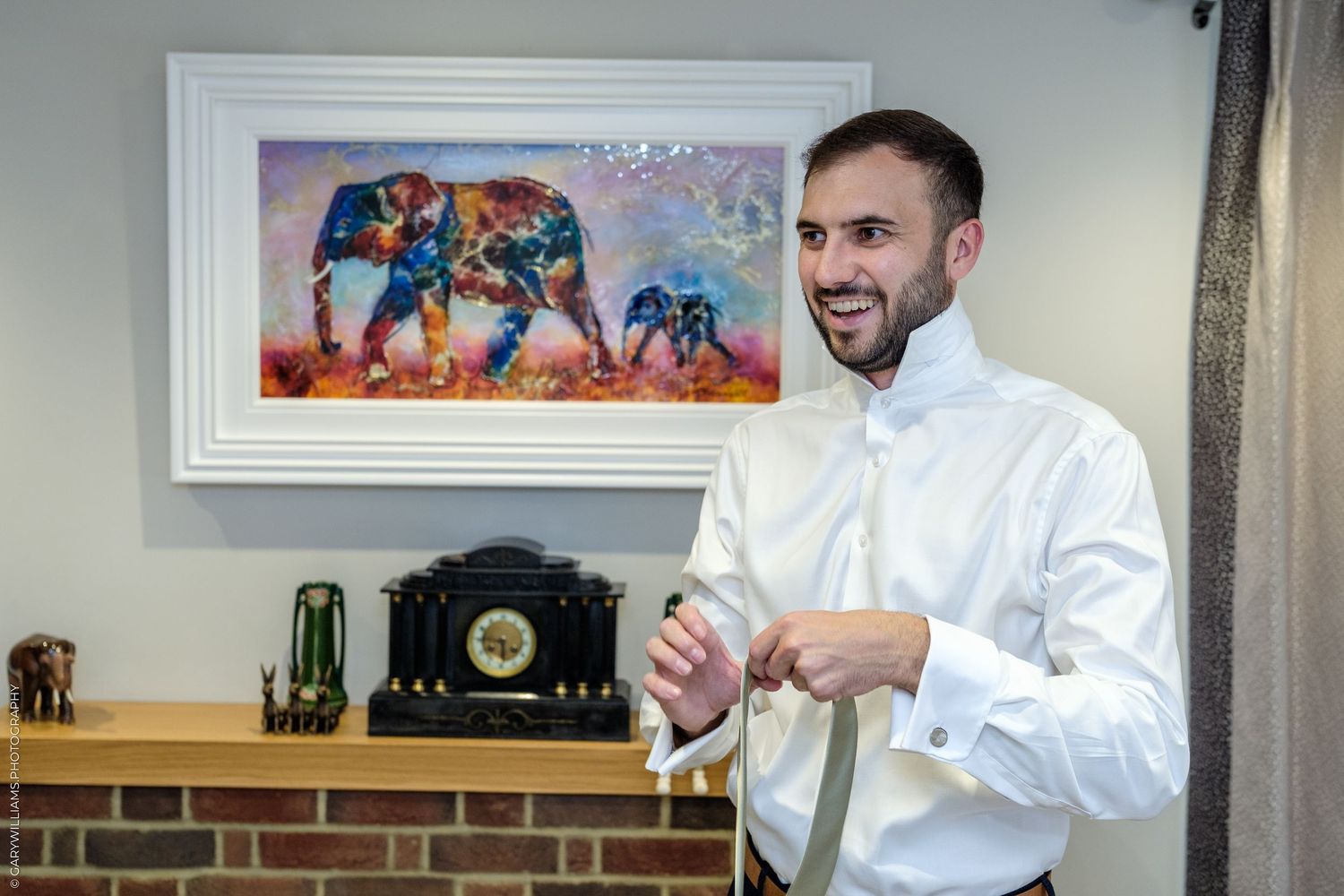 A smiling groom in a white dress shirt adjusting his tie near a fireplace with colorful elephant artwork on the wall.