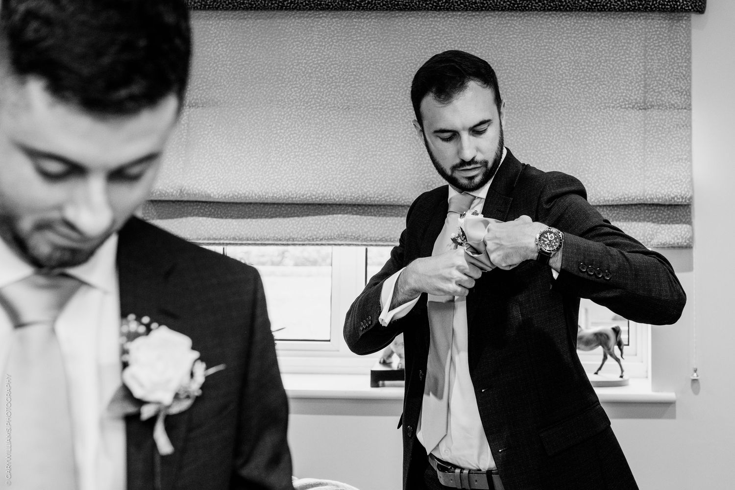 Two groomsmen adjusting their ties and boutonnieres in black and white wedding preparation photo.