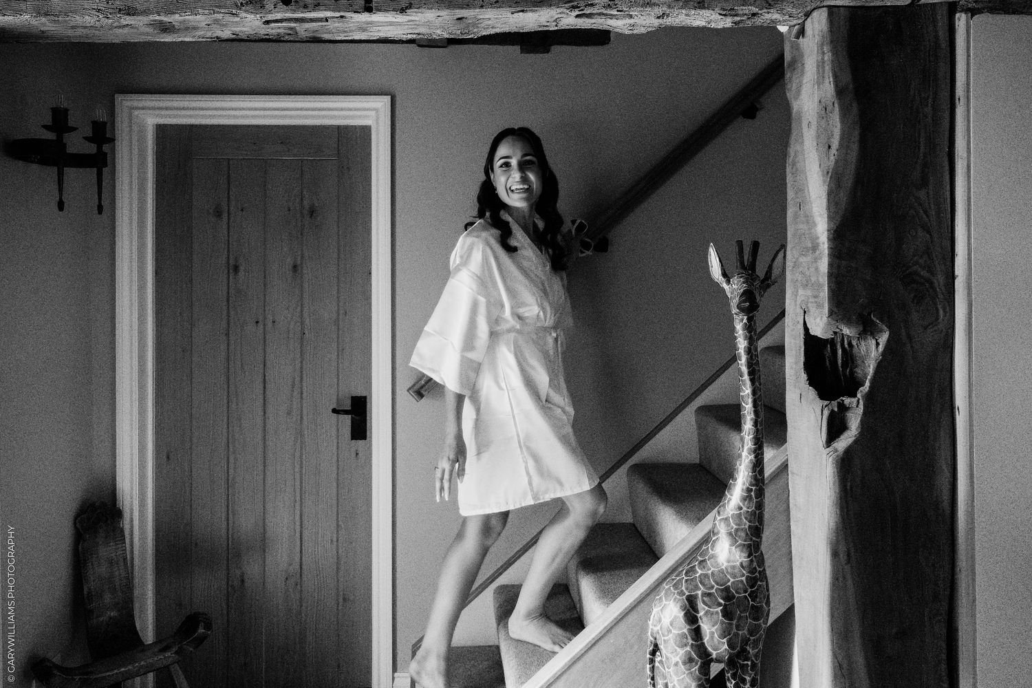 Black and white photo of a bride in a white robe standing on stairs in a rustic venue.