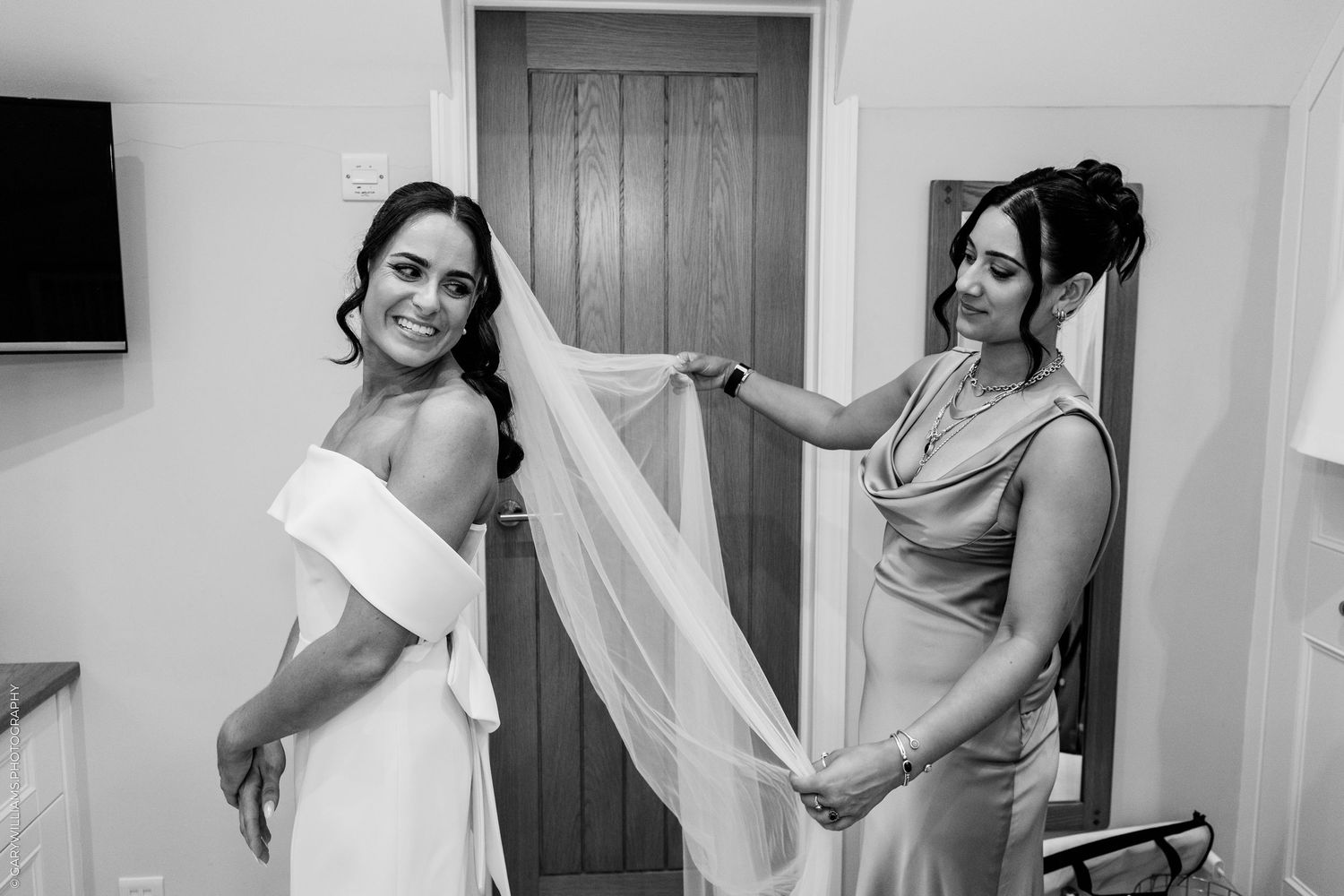Black and white photo of a bride in a white gown as her bridesmaid adjusts her veil before the wedding ceremony.