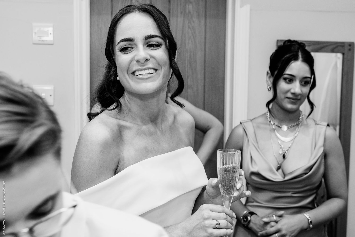 Emotional black and white photo of a bride smiling with bridesmaids holding champagne glasses during wedding preparations.