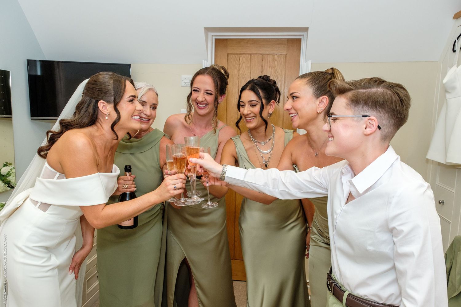 Bride and bridesmaids in sage green dresses toasting with champagne glasses at a wedding celebration indoors.
