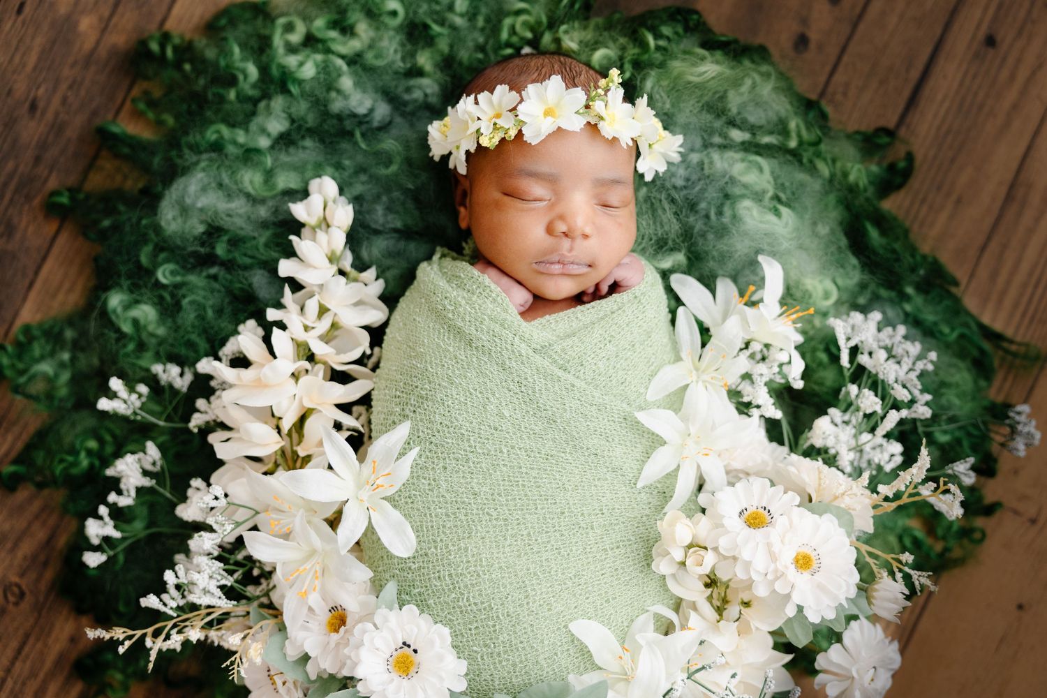 Newborn baby wrapped in green swaddle with floral crown, surrounded by white flowers on lush green backdrop.