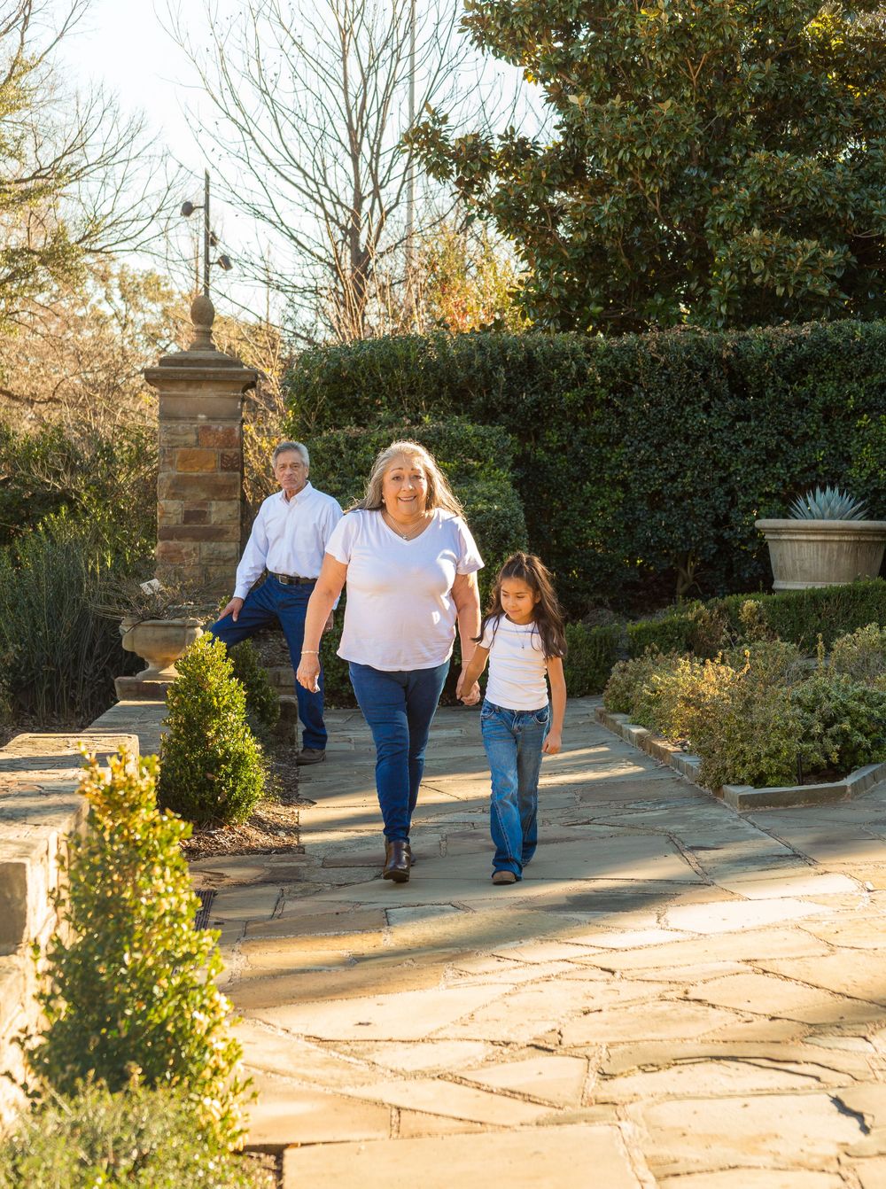 Three generations walk together on a stone garden path surrounded by lush hedges and trees on a sunny day.
