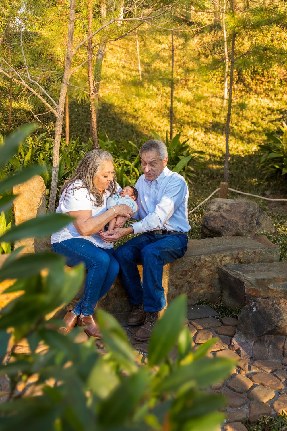 Grandparents sitting on stone steps in a sunlit garden, lovingly cradling a newborn baby together.