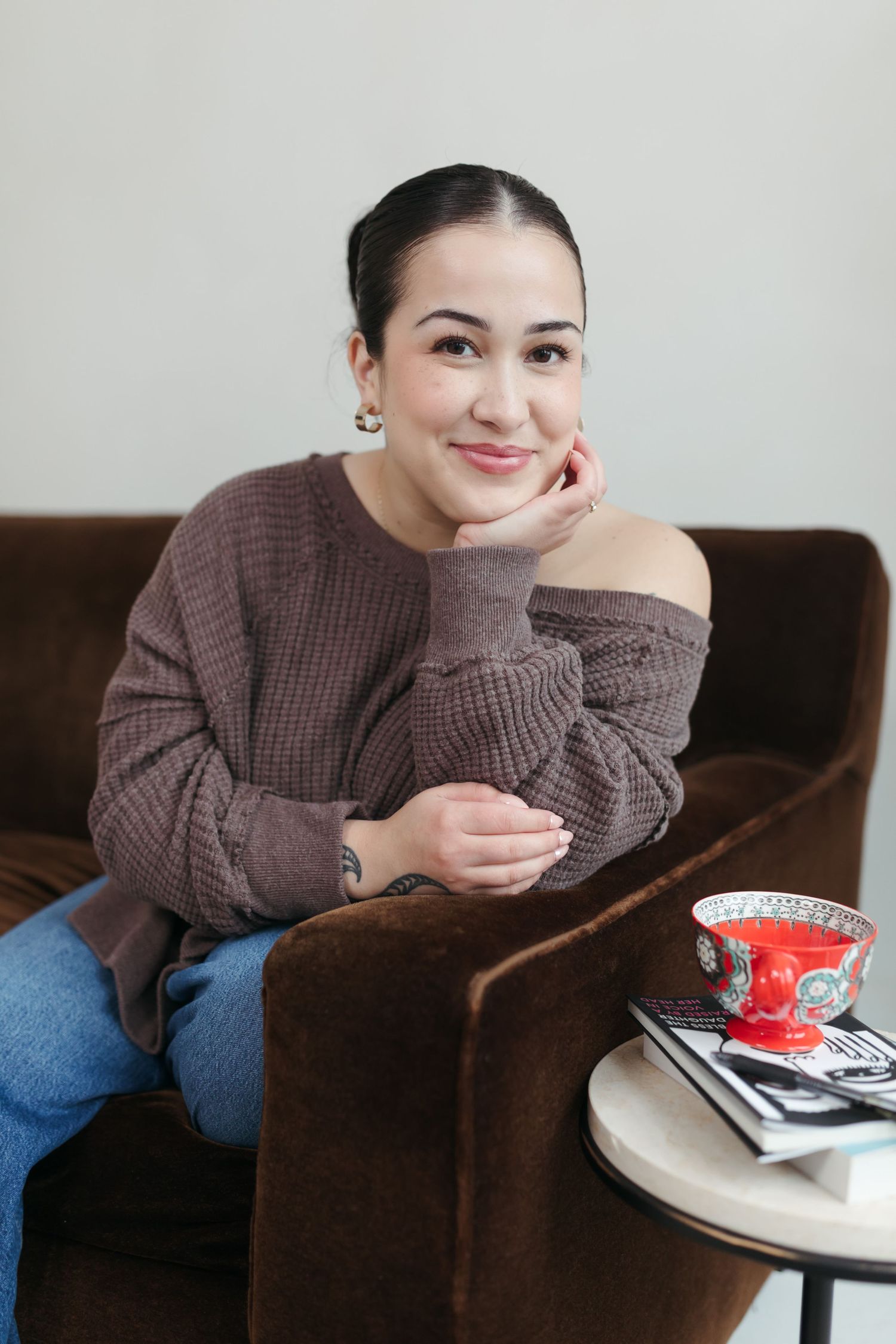 Woman in brown sweater sitting in velvet chair, smiling with hand on chin, red teacup on side table.