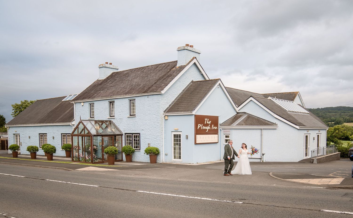 Bride and groom pose outside The Plough Inn, a charming white country hotel surrounded by lush greenery.