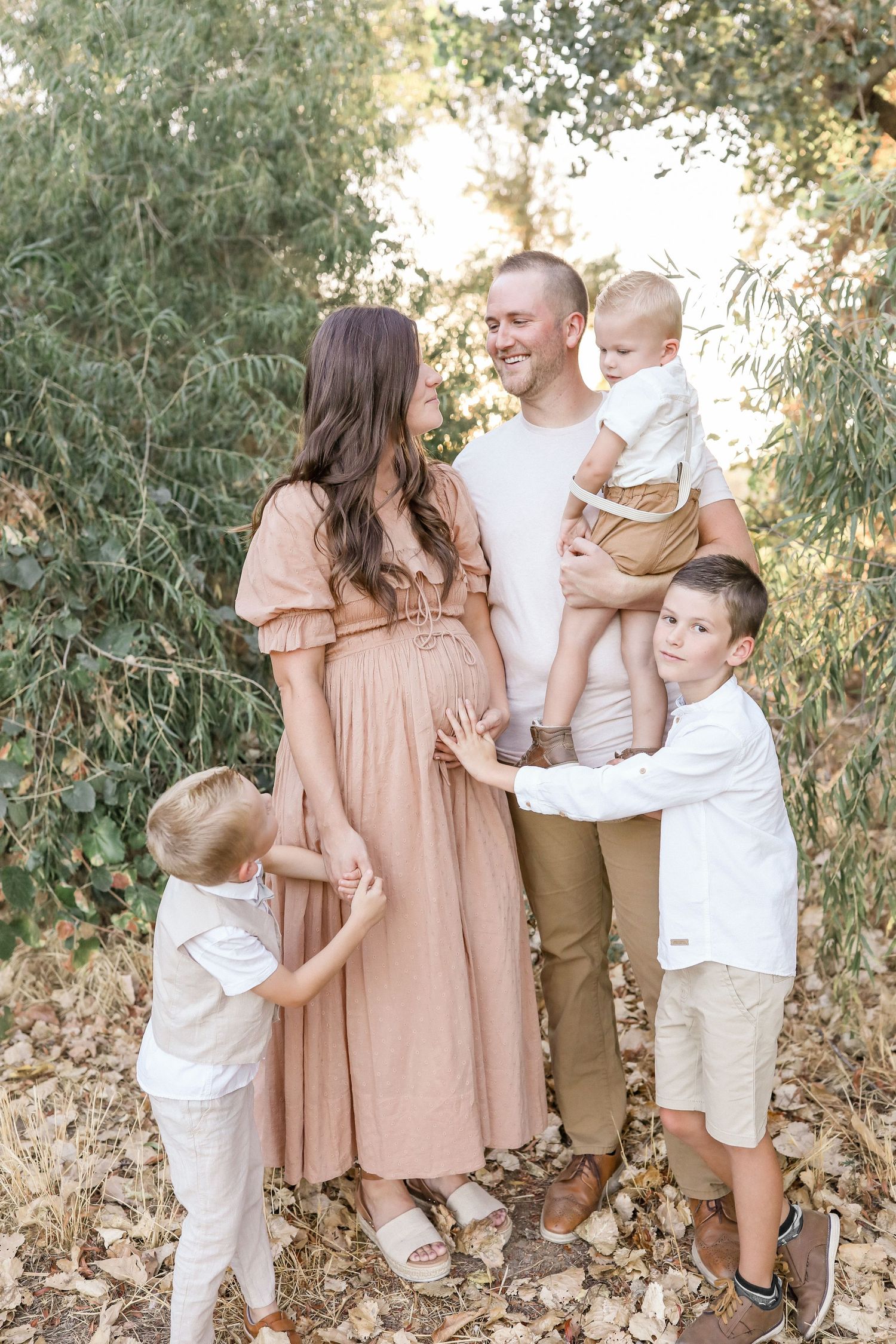 Pregnant woman in pink dress with husband and two boys touching baby bump outdoors.