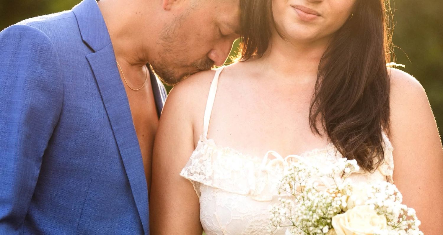 Romantic couple sitting together in a sunlit meadow, man in blue suit embracing woman in white lace dress.