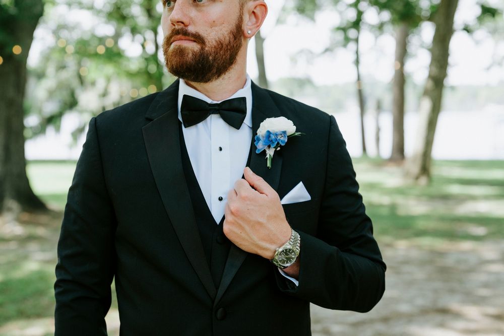 Groom in black tuxedo with bow tie adjusting blue boutonniere and white pocket square outdoors.