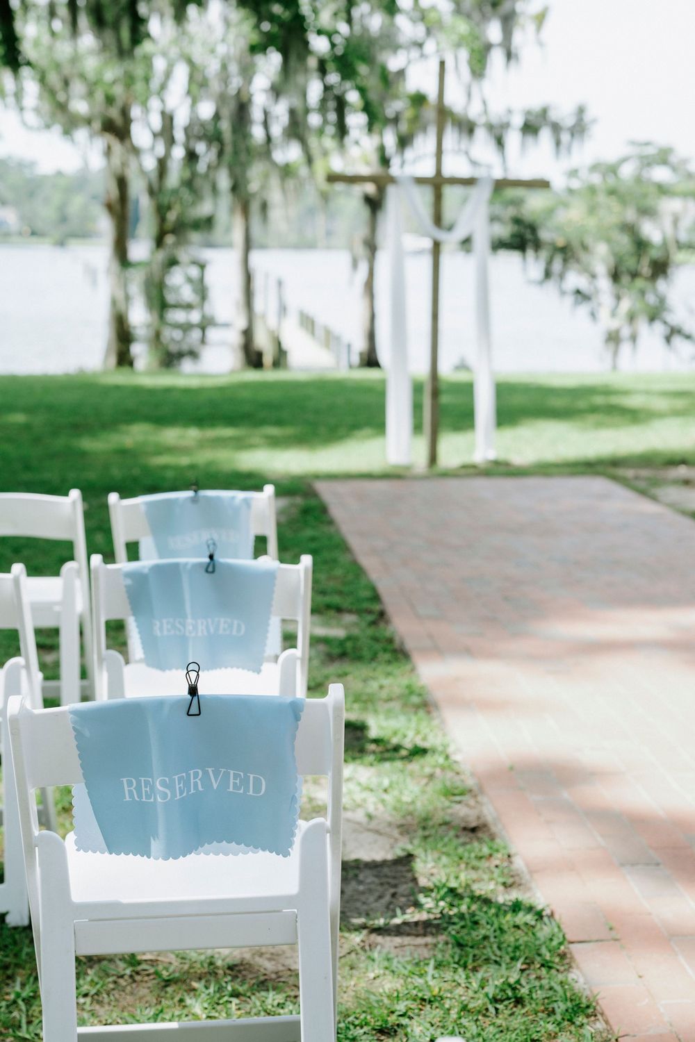 White folding chairs with light blue reserved signs line a brick aisle leading to a wooden cross altar by a lakeside.