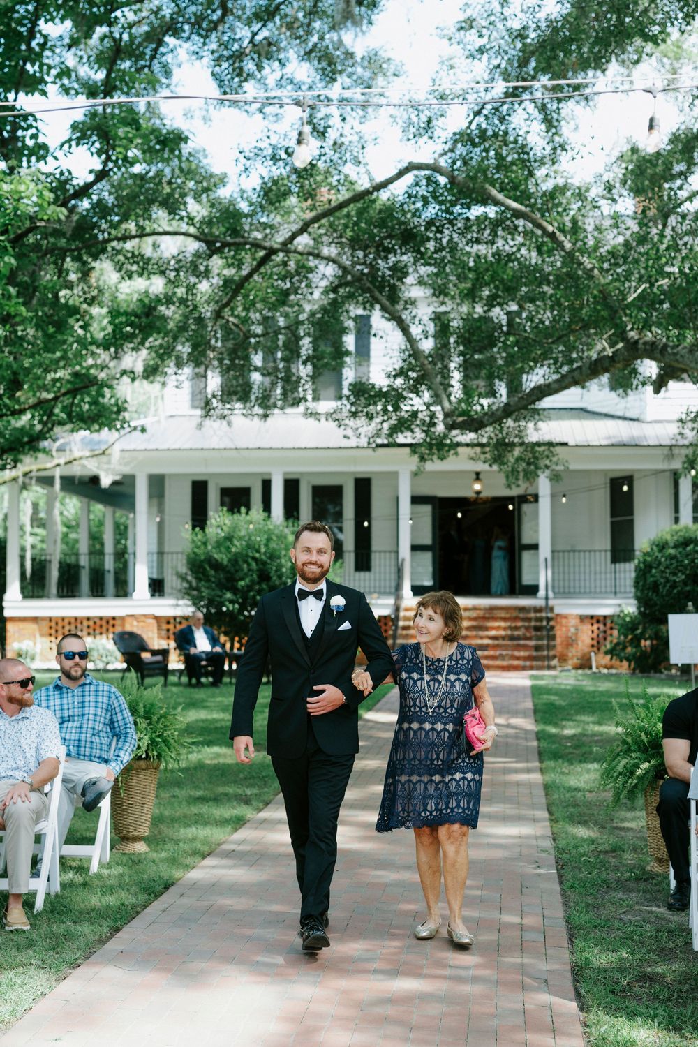 Groom in black tuxedo walks down outdoor wedding aisle with his mother beside a white Southern mansion.
