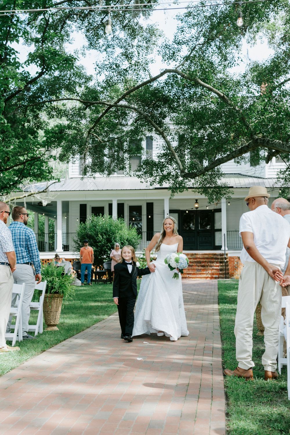Bride walks down outdoor aisle with young boy in tuxedo, white colonial venue, lush green trees, wedding guests seated.