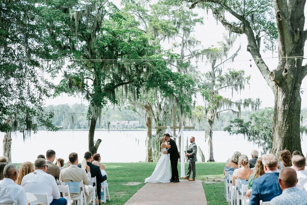 Outdoor wedding ceremony under moss-draped oak trees by a serene lake, with guests seated on white chairs.