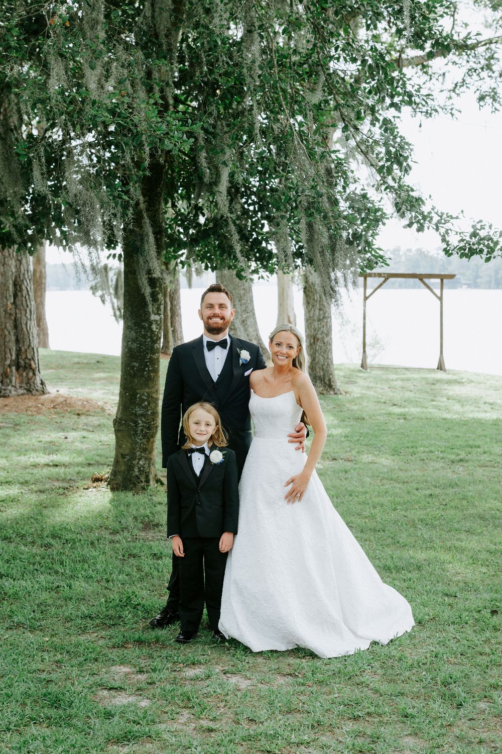 Bride and groom in wedding attire pose with young boy in tuxedo under mossy trees near lake.