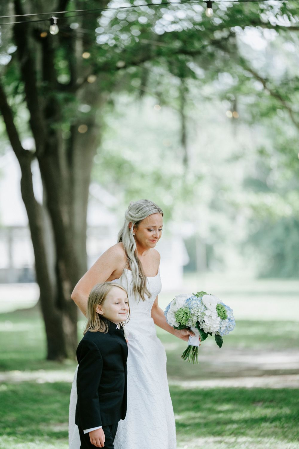 Smiling bride in white strapless gown holds blue and white bouquet while standing with young boy.
