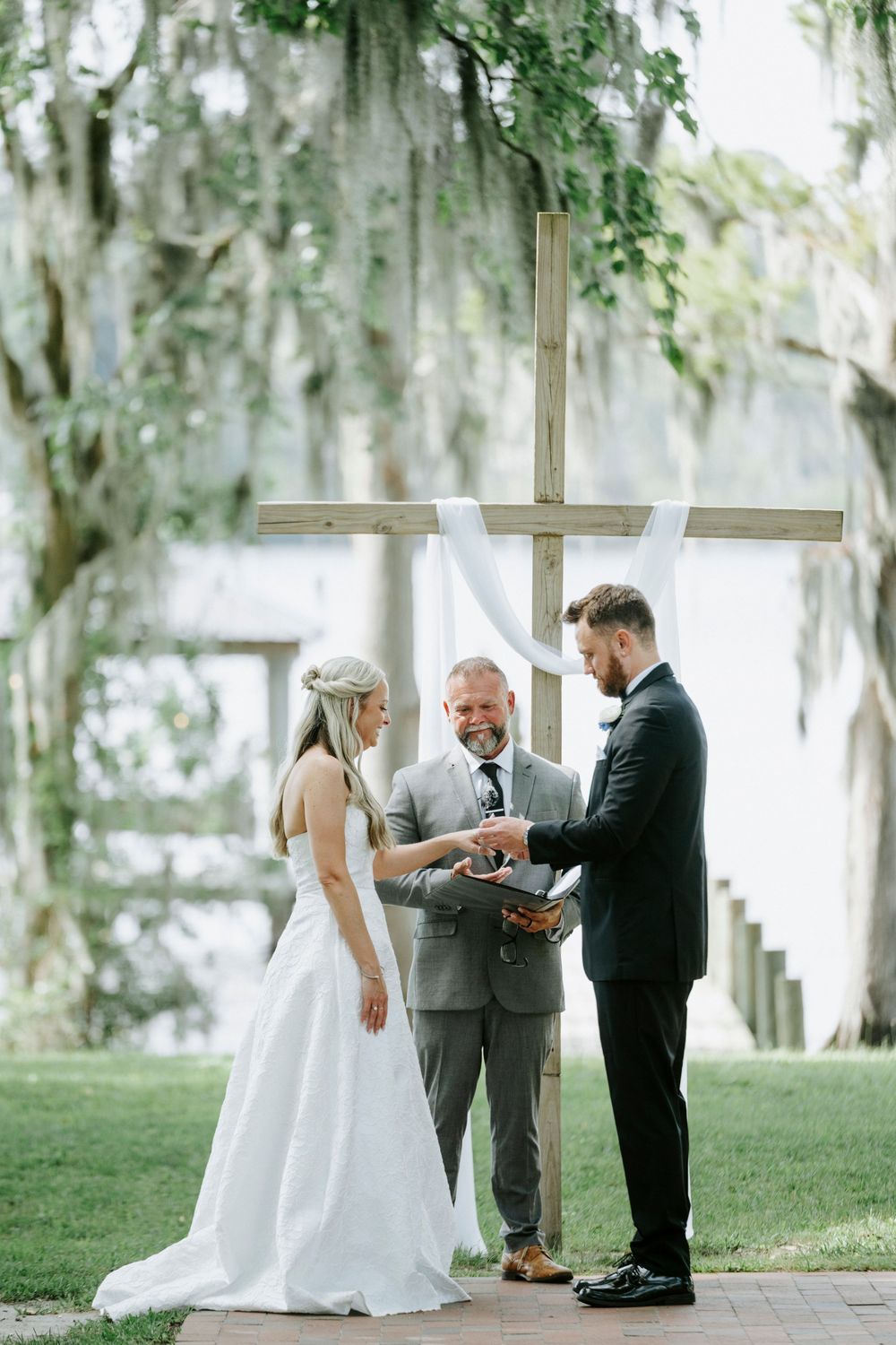 Bride and groom exchange vows outdoors before a wooden cross draped in white fabric, surrounded by moss-covered trees.