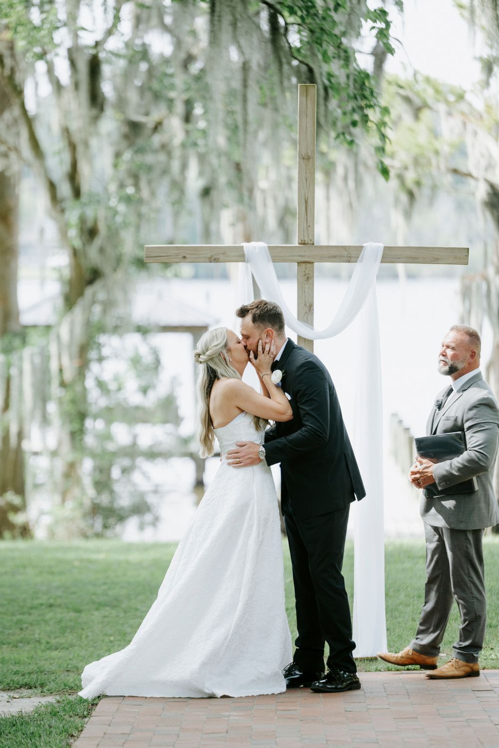 Bride and groom share first kiss at outdoor lakeside ceremony before wooden cross altar with white draping.
