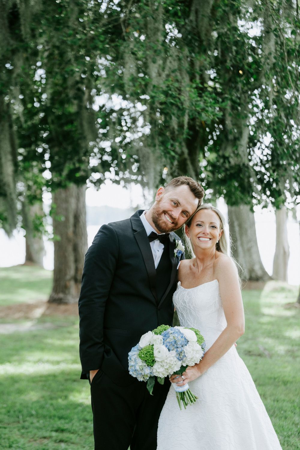 Smiling bride and groom pose under lush green trees, bride holding a bouquet of blue hydrangeas and white flowers.