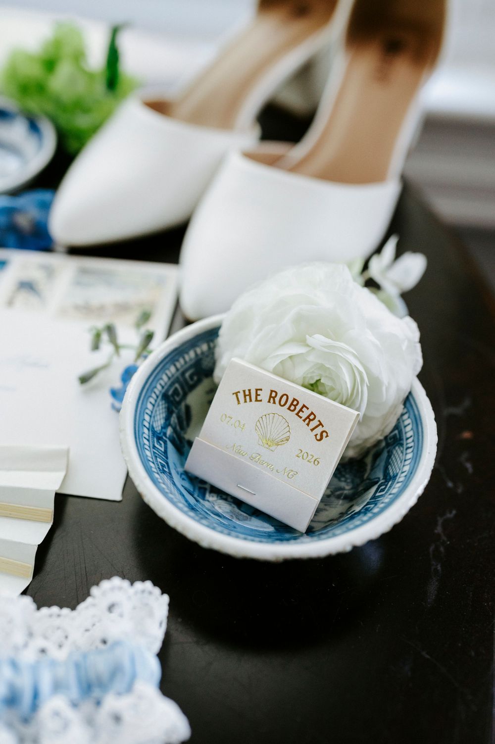 Wedding details flat lay with white bridal heels, ranunculus flower, and The Roberts monogram matchbox in blue bowl.