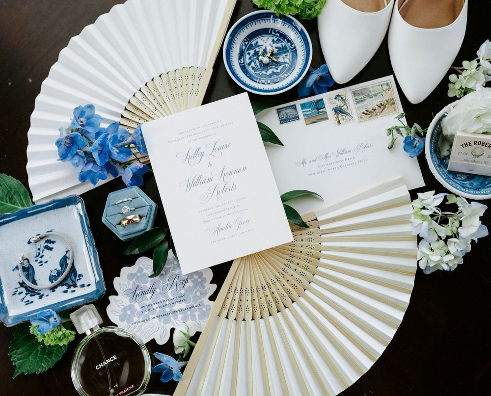Elegant wedding flat lay with white paper fans, blue floral accents, invitation suite, rings, and Chanel perfume on dark background.