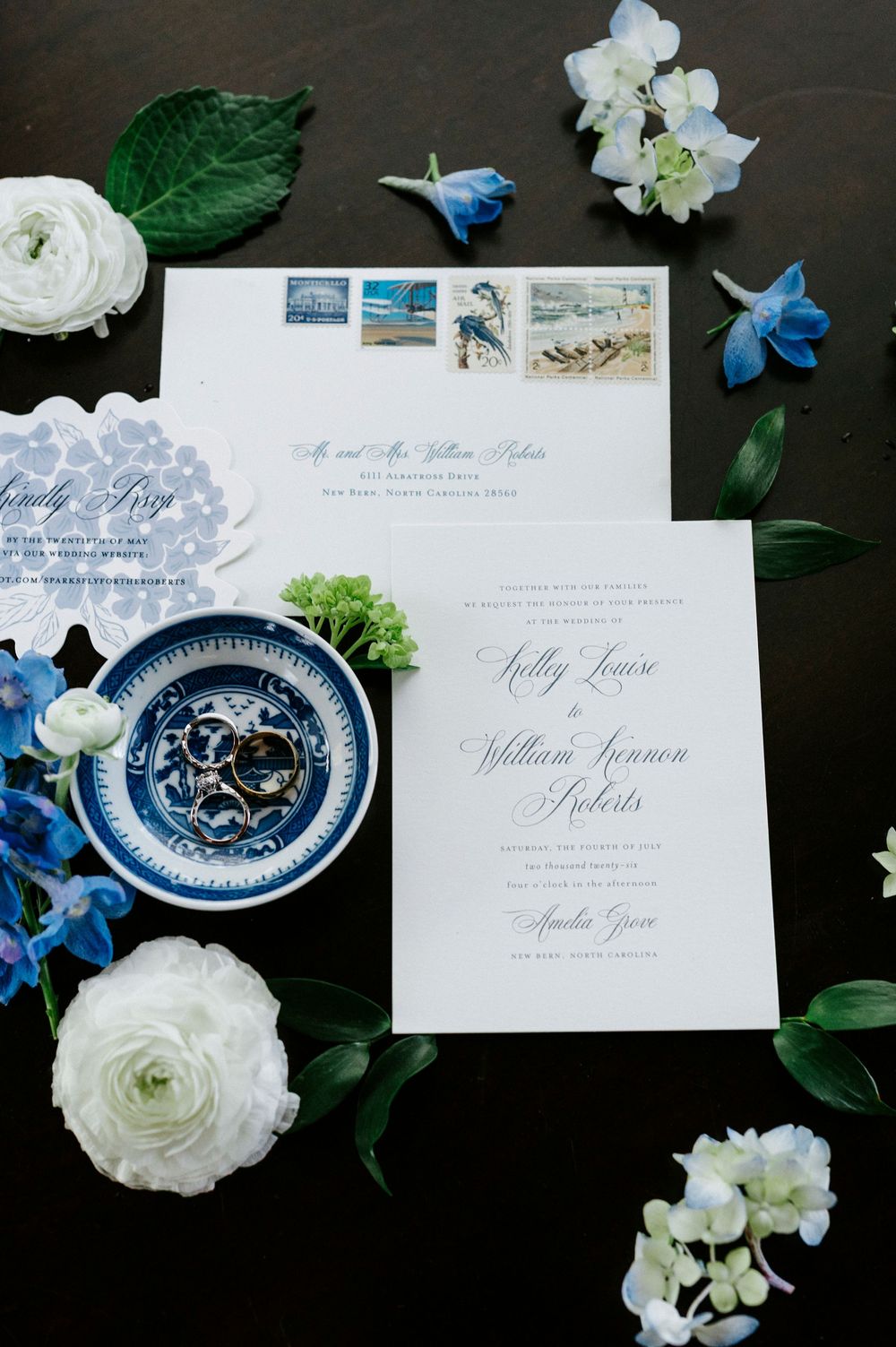 Elegant blue and white wedding invitation suite surrounded by fresh flowers and greenery on dark background.