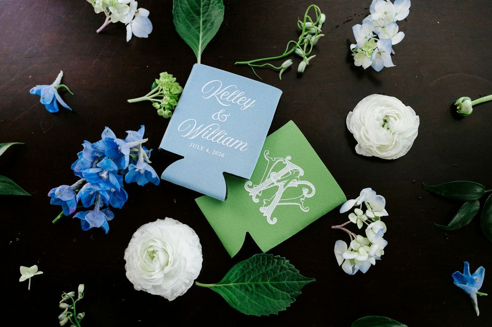 Blue and green personalized wedding koozies for Kaitei and William surrounded by white ranunculus and blue delphinium flowers.