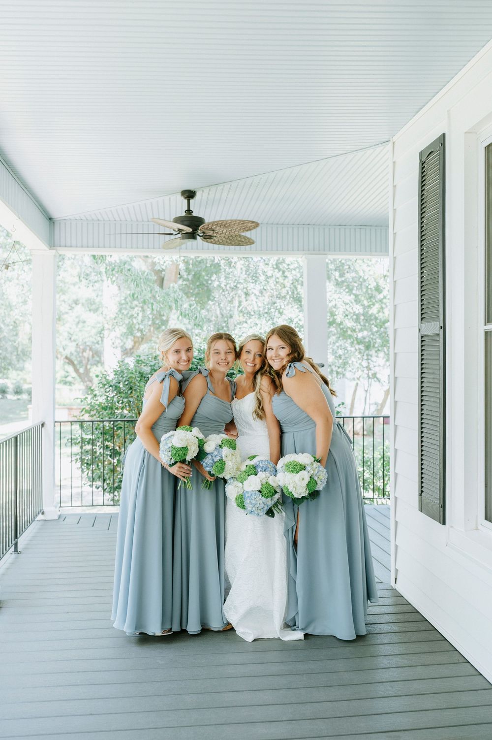 Bride and two bridesmaids in dusty blue gowns holding white hydrangea bouquets on a white porch.