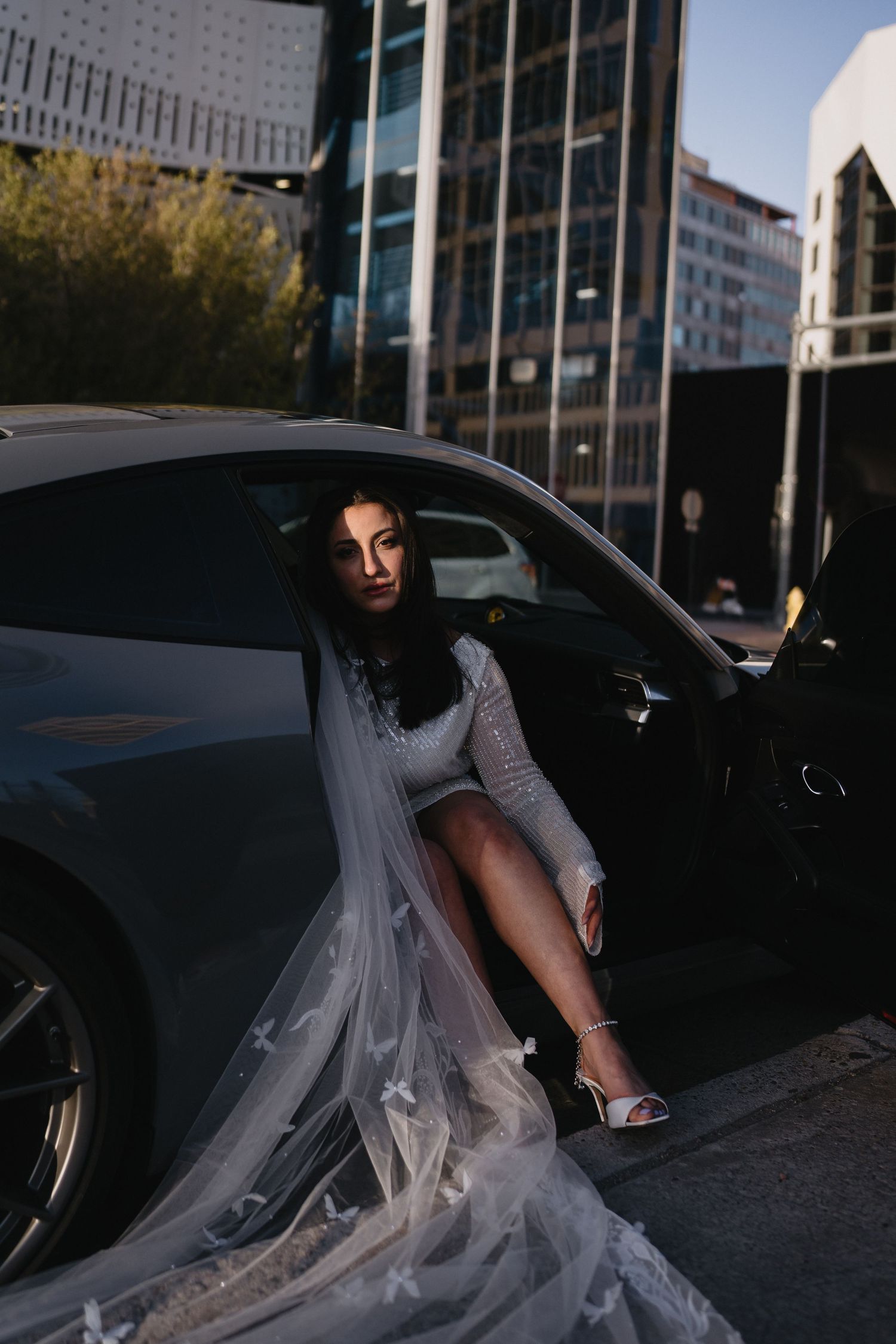Bride in white lace dress and veil sitting in a gray sports car door in an urban environment in Downtown Albuquerque
