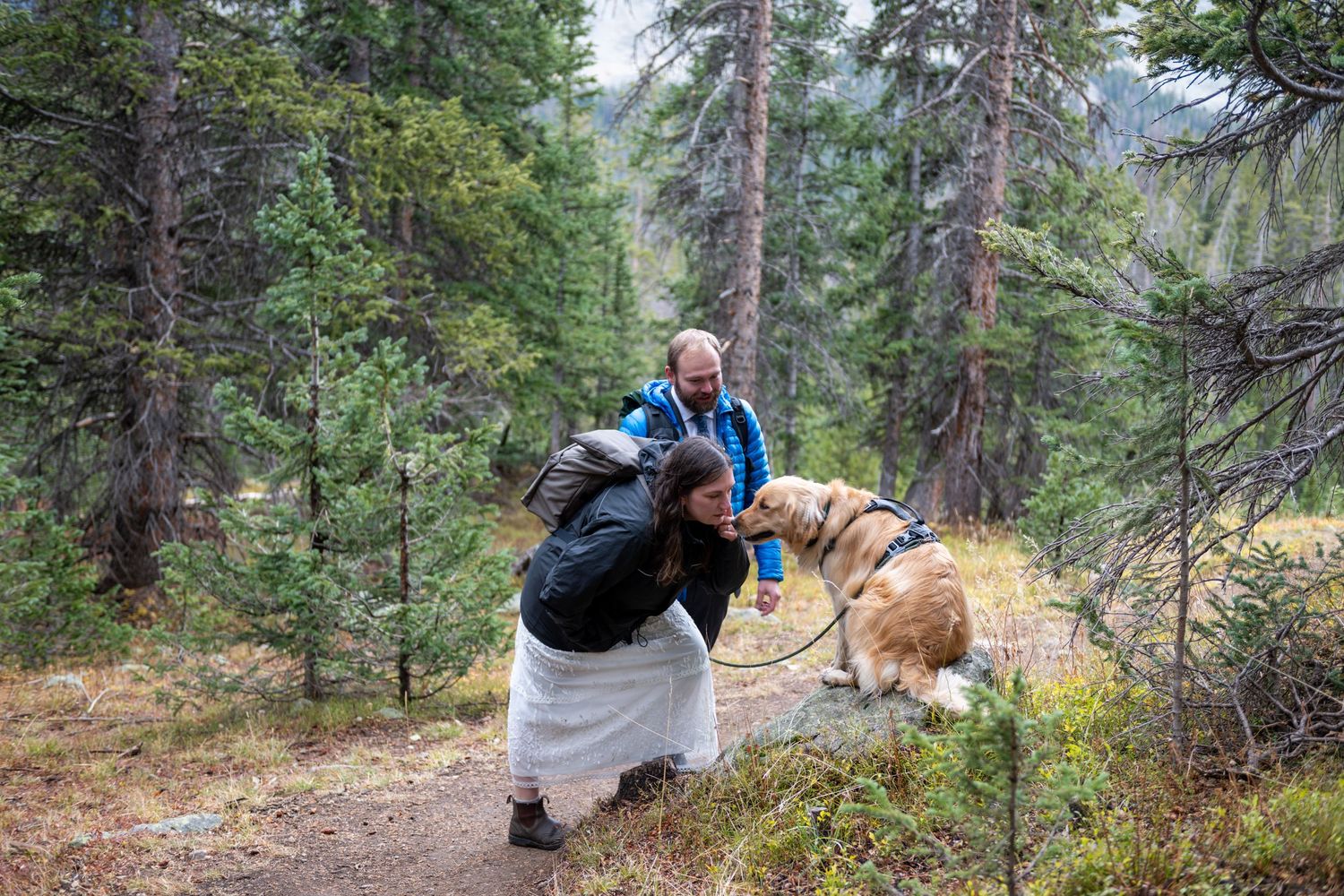 An eloping couple stop on the hiking trail to give their dog kisses. They are wearing rain jackets and back packs and wedding attire.