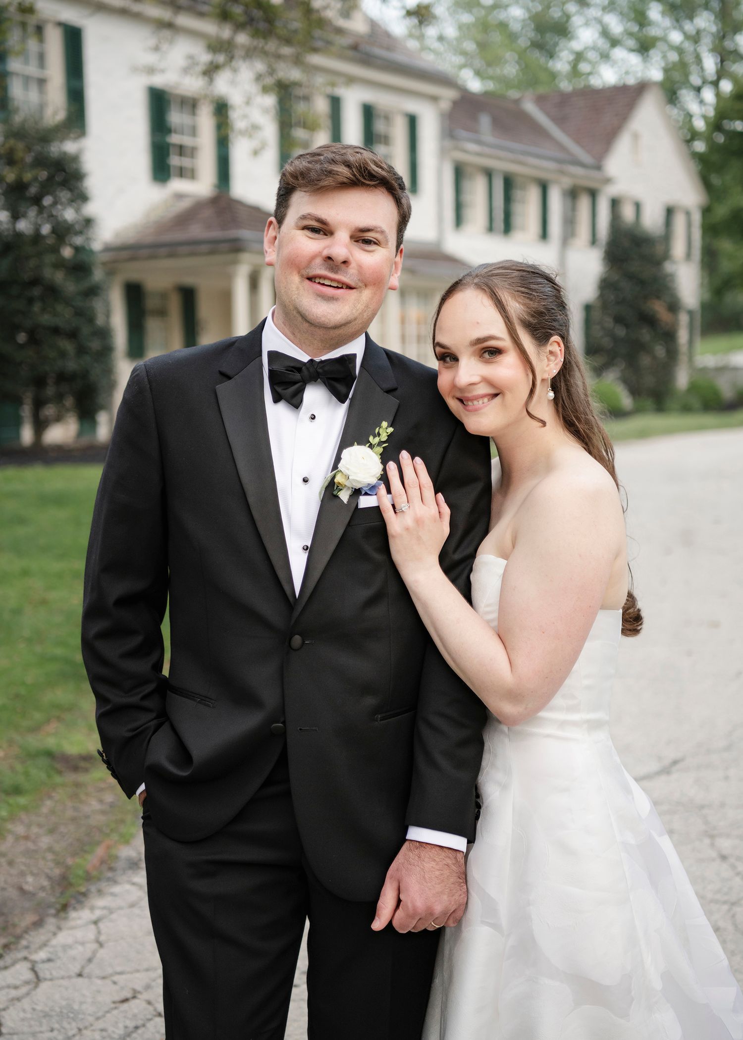 Bride and groom pose together in formal wedding attire outside an elegant white manor house surrounded by spring trees.
