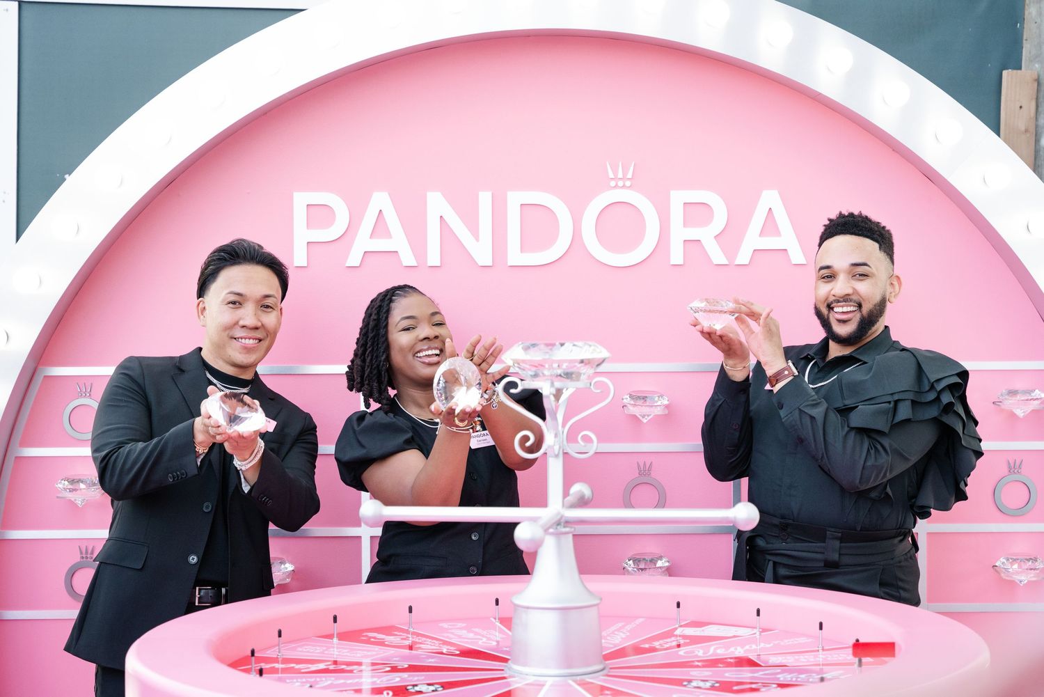 Three people smile holding Pandora jewelry in front of a pink Pandora branded display at a promotional event.