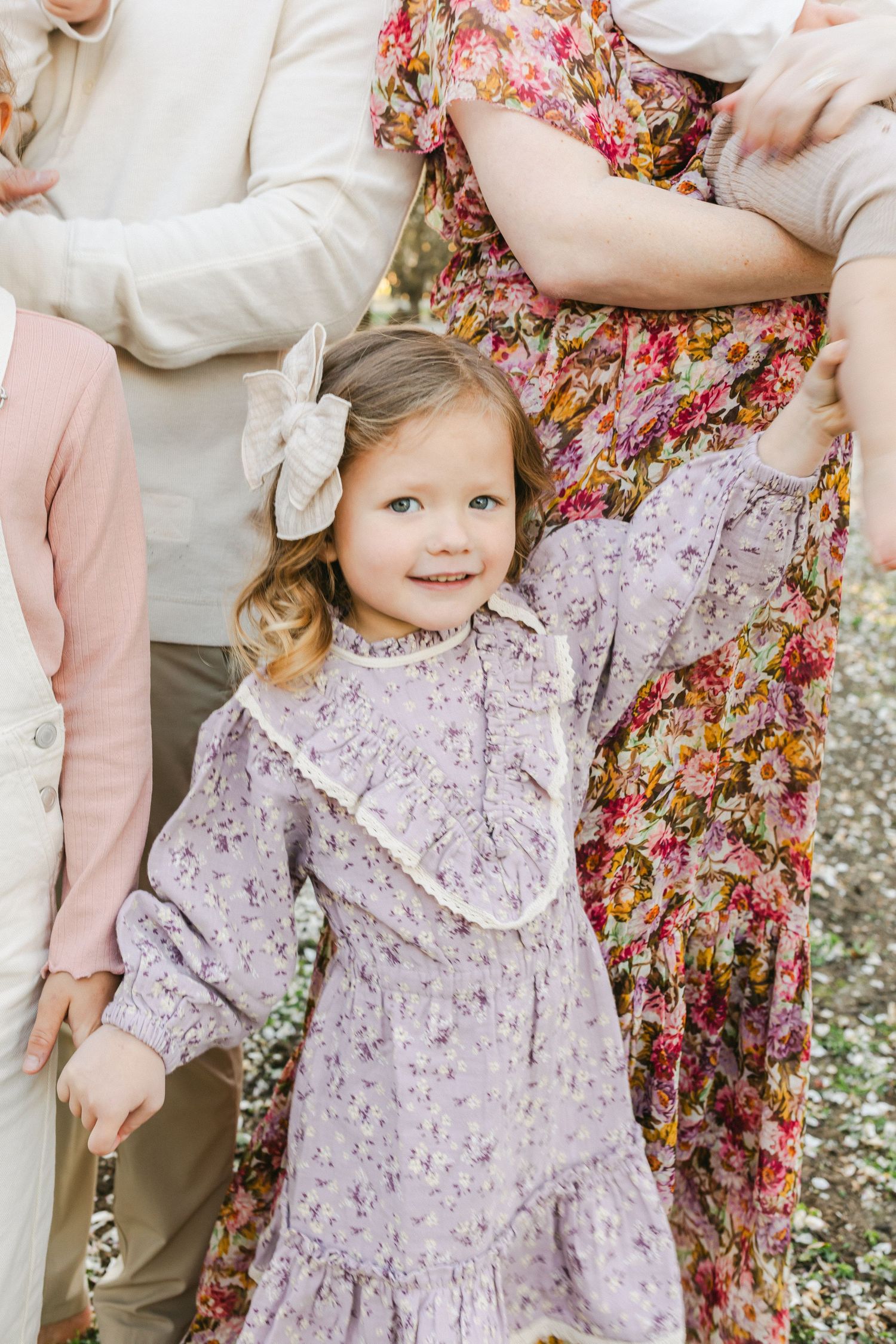 A joyful toddler girl in a floral dress and bow hugs an adult's legs outdoors among white flowers.