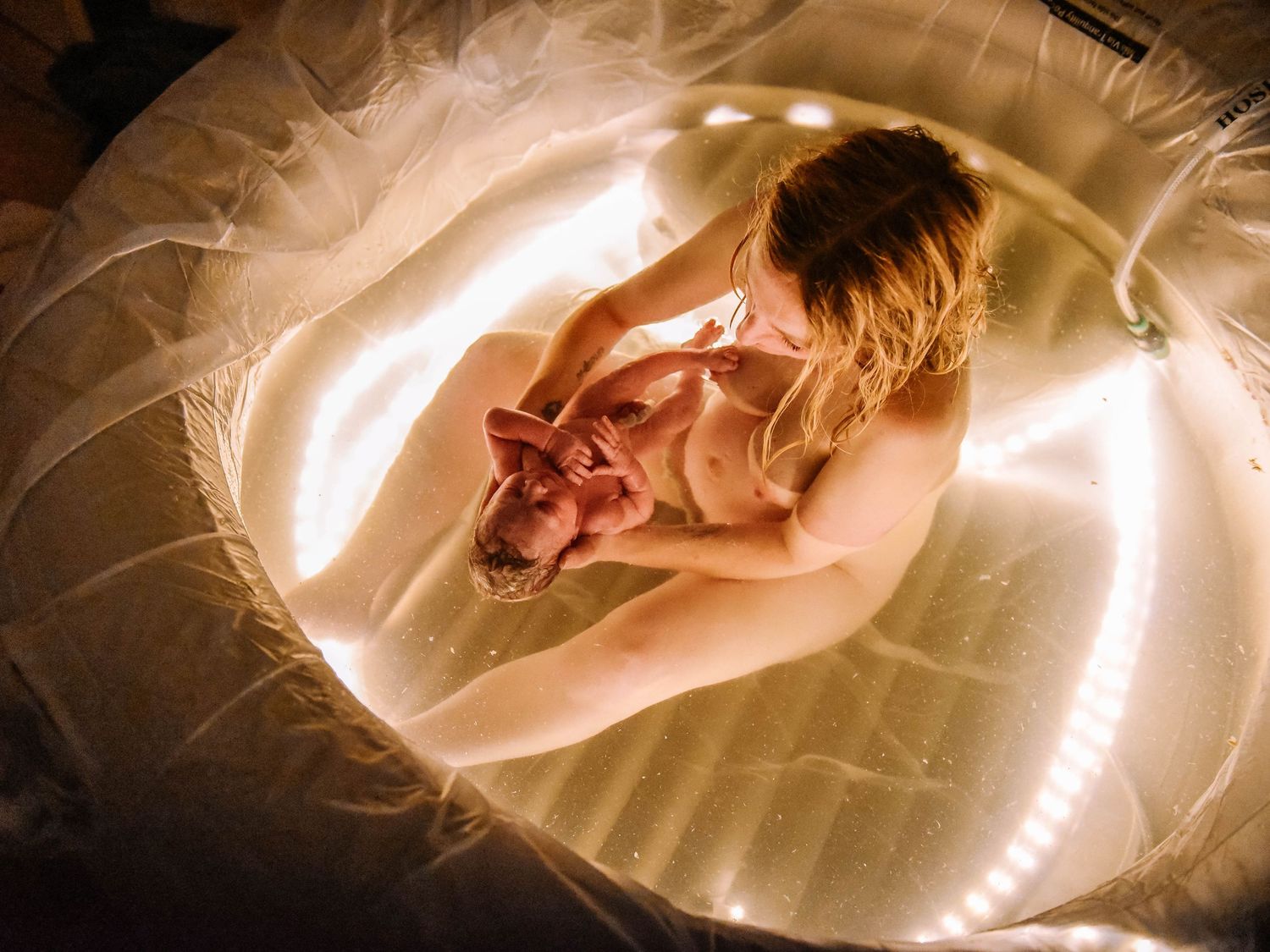 Aerial view of a mother cradling her newborn in a glowing birthing pool.