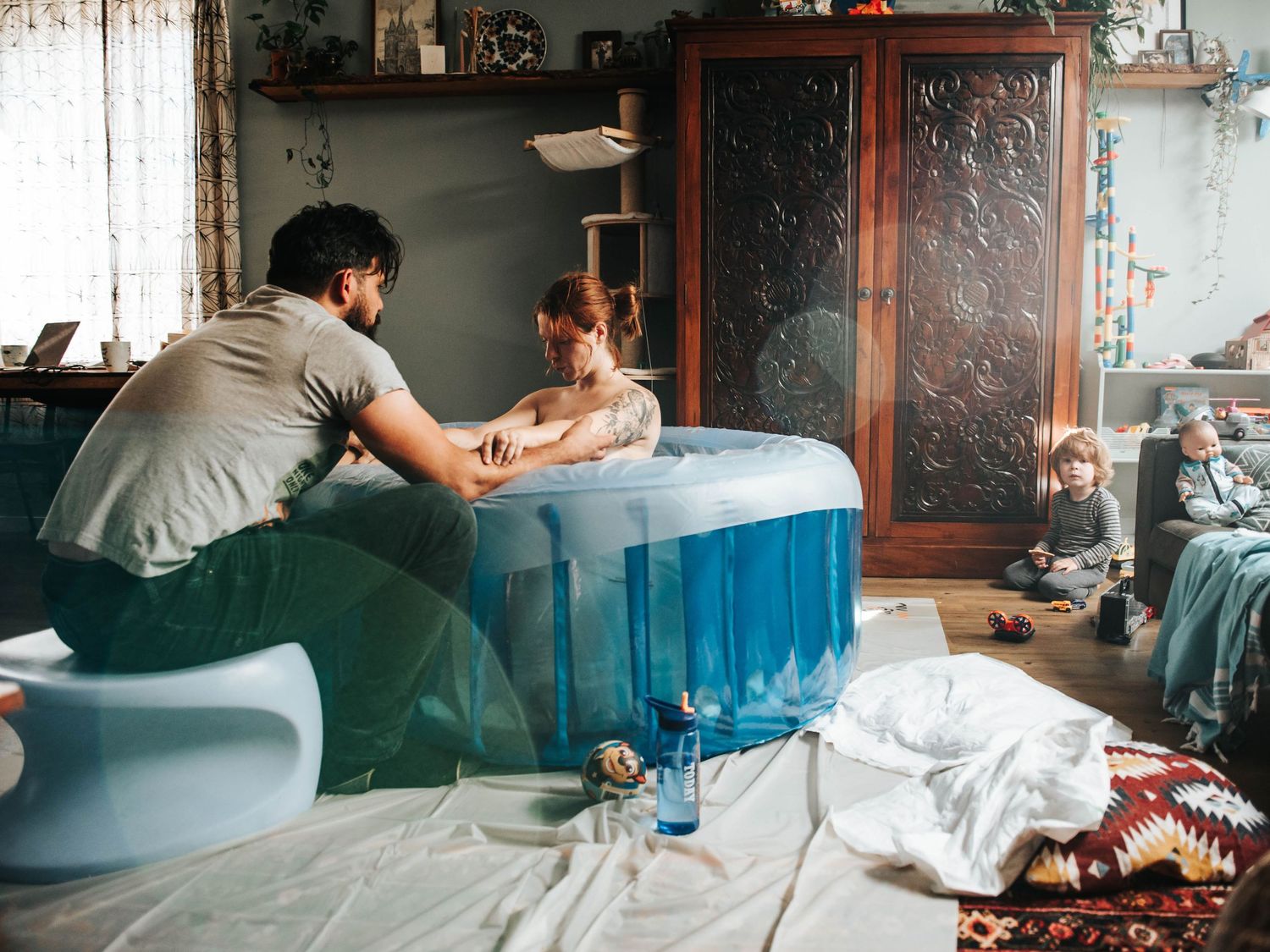 Man assists woman in a blue inflatable birth pool at home, with a child playing nearby in a cozy living room.