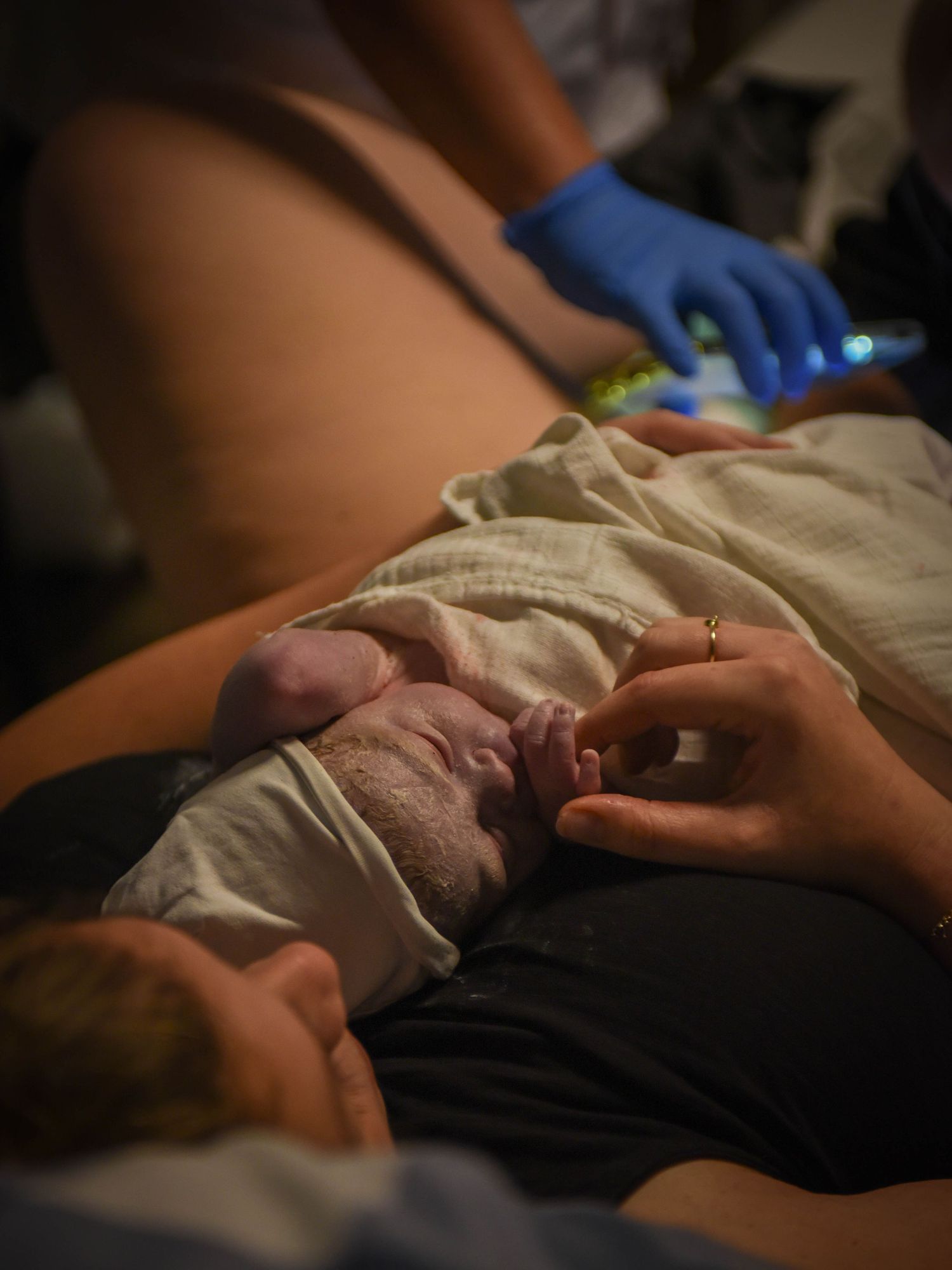Healthcare worker in blue gloves assists as newborn lies on mother's chest during postpartum care after delivery.
