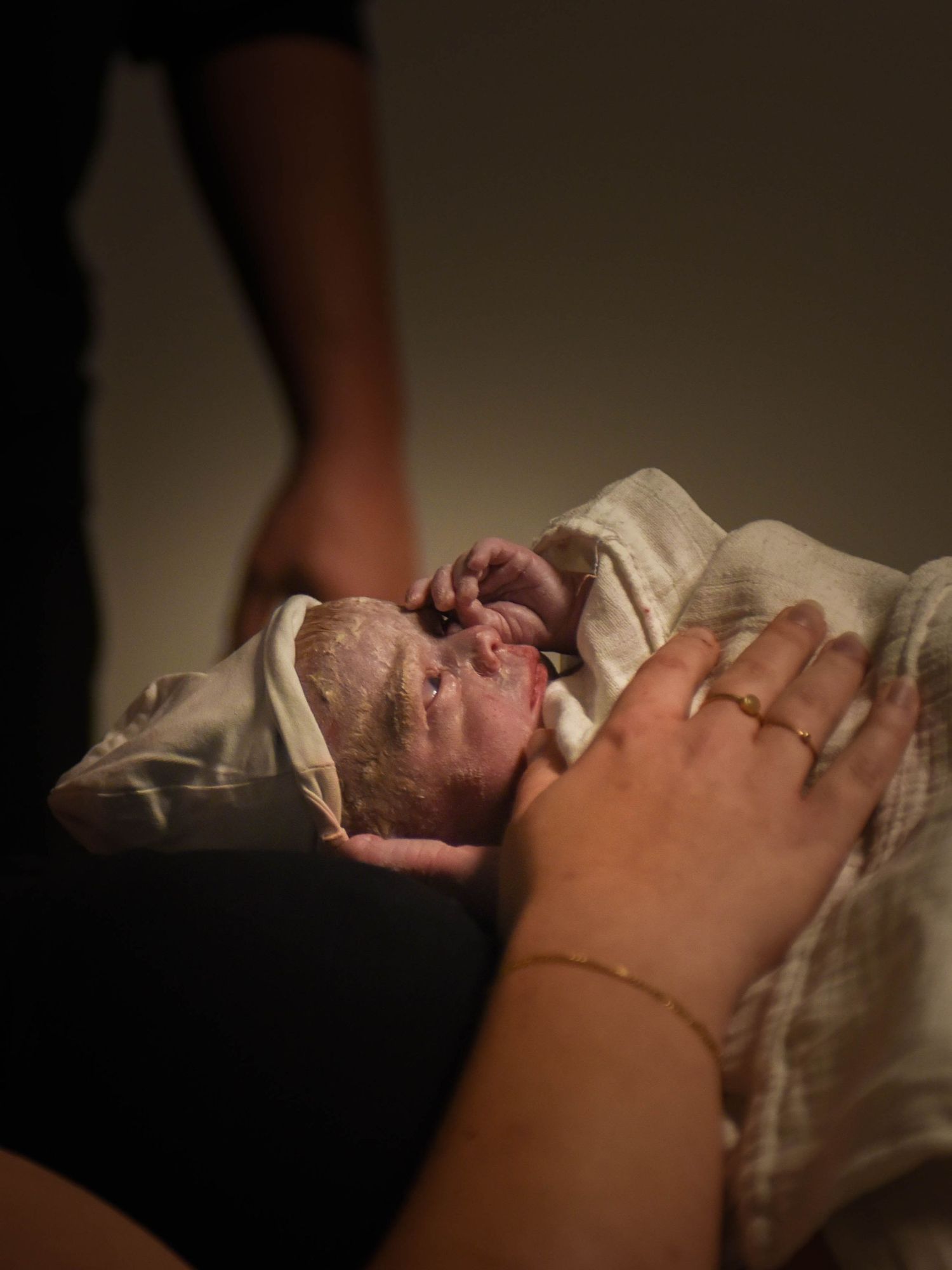 A newborn baby wrapped in white blanket being held by a mother wearing a gold bracelet and ring in soft dim light.