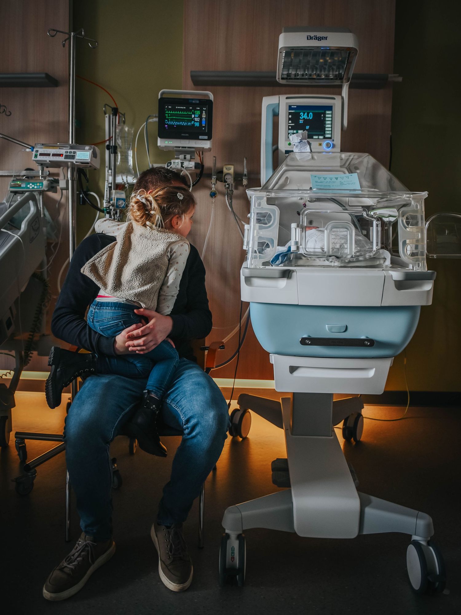 Parent sitting beside a neonatal incubator in a NICU, monitoring their newborn with medical equipment nearby.