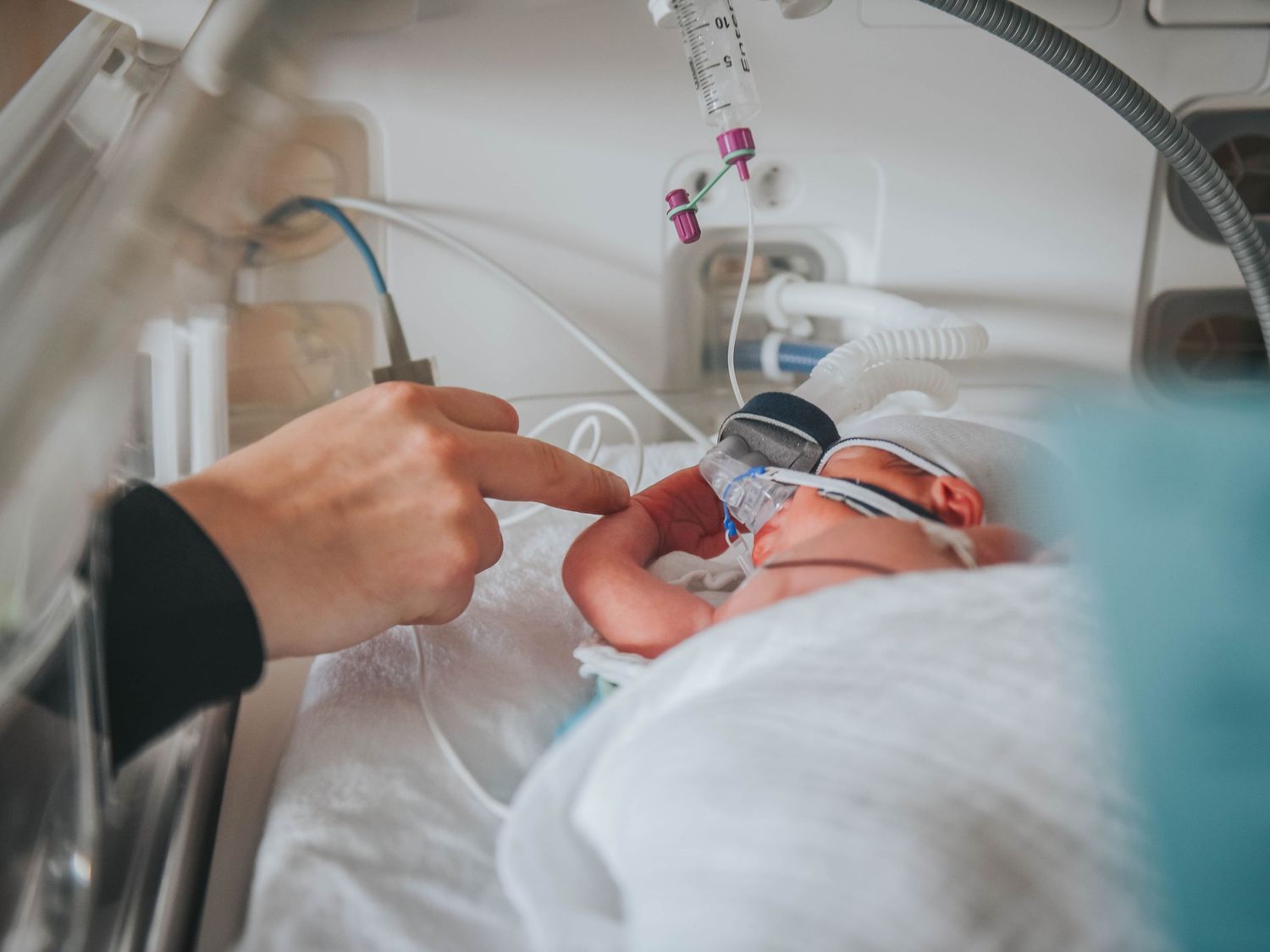 Premature newborn baby in hospital incubator with medical equipment, as a parent gently touches the infant's hand.