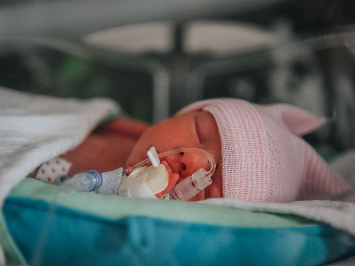 Premature newborn baby sleeping in NICU incubator with medical tubes and pink hat in hospital.