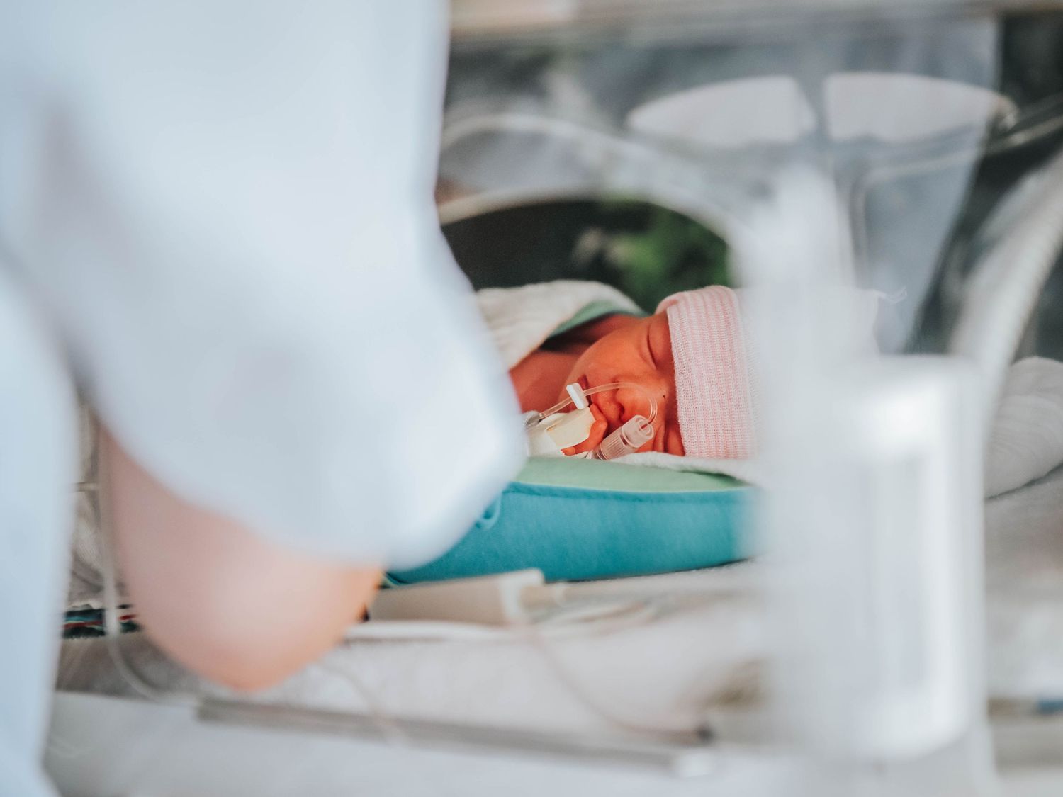 Premature newborn baby resting in a hospital incubator with medical equipment, attended by a nurse in white scrubs.