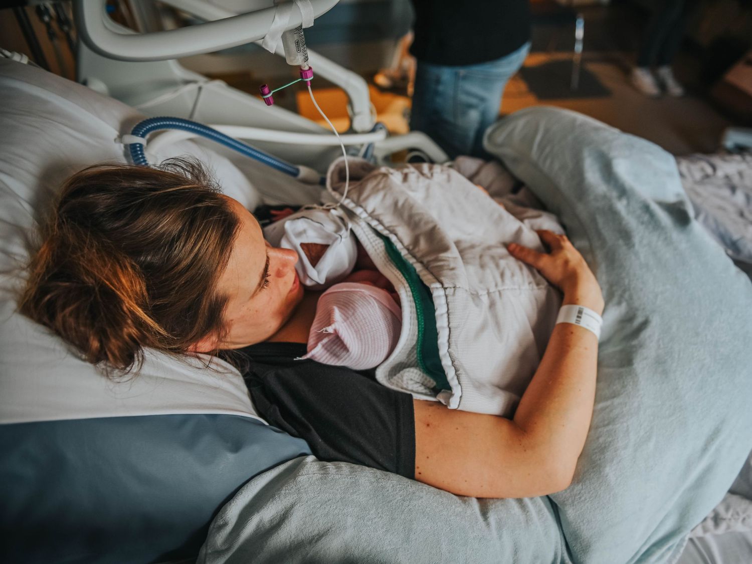 A new mother lying in a hospital bed tenderly holding her newborn baby with medical equipment visible nearby.