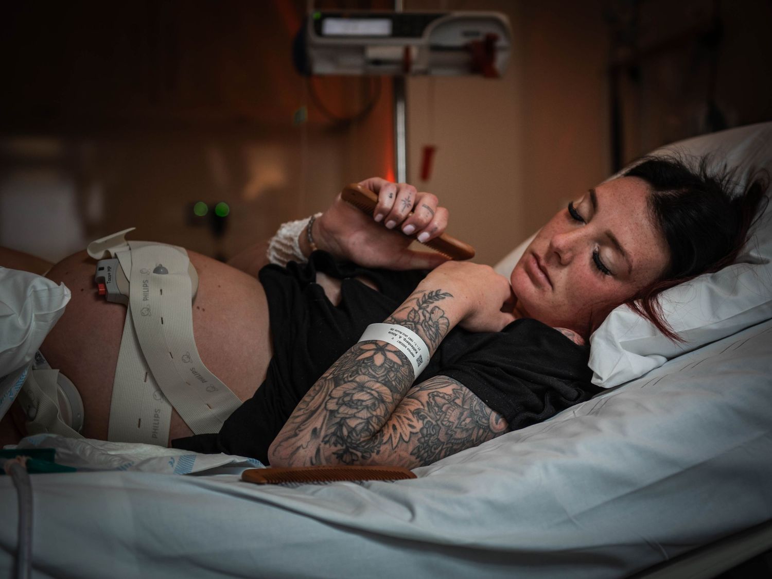 Tattooed woman resting in hospital bed, wearing black shirt and patient wristband, looking at phone.
