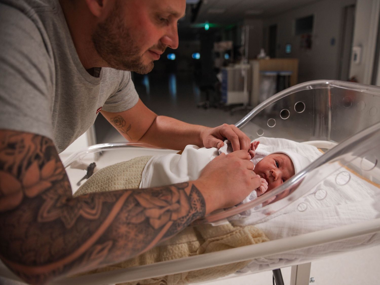 Tattooed father gazing lovingly at newborn baby in hospital bassinet shortly after birth.