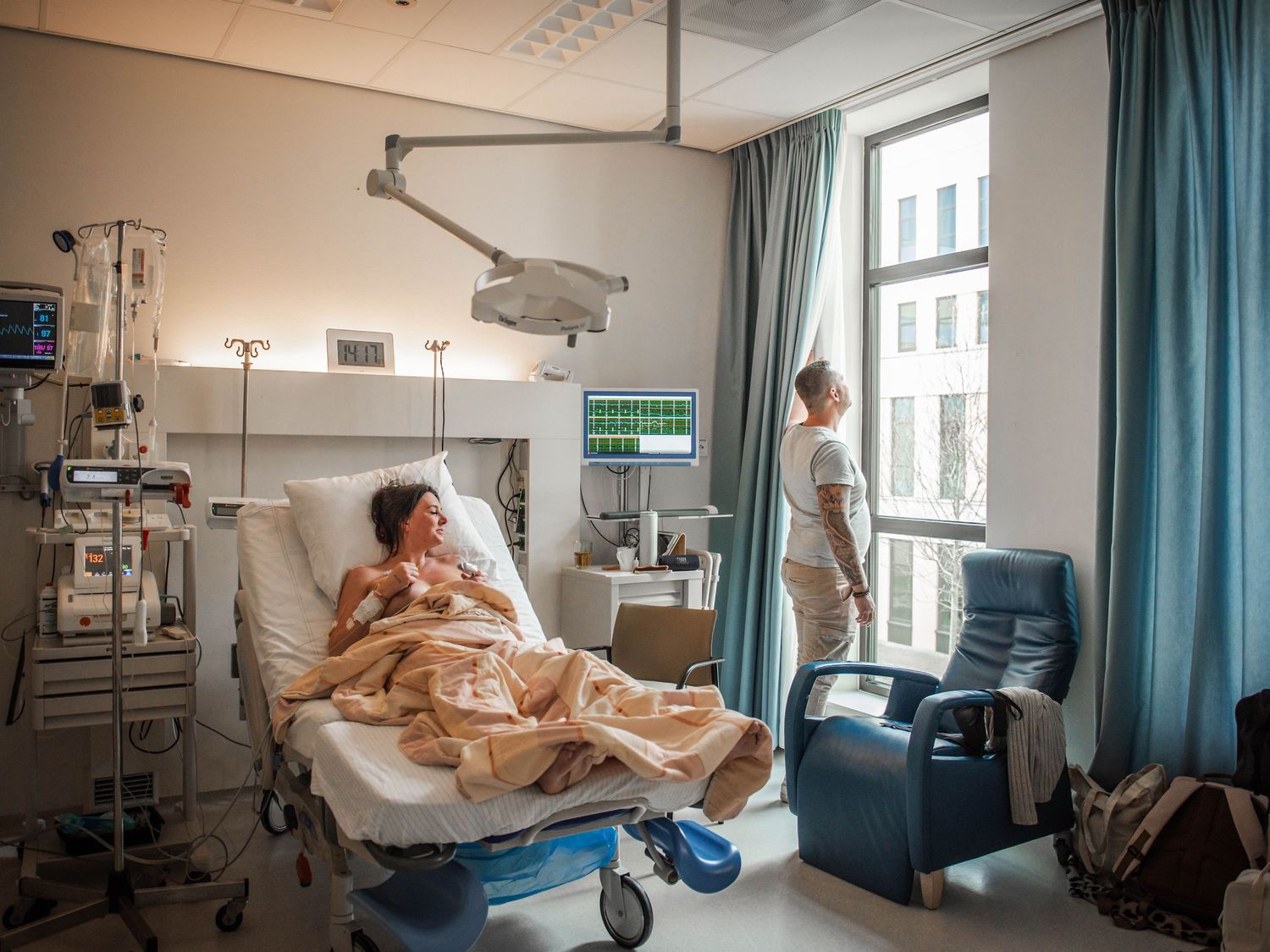 Patient resting in hospital bed while nurse stands by window in modern private hospital room.