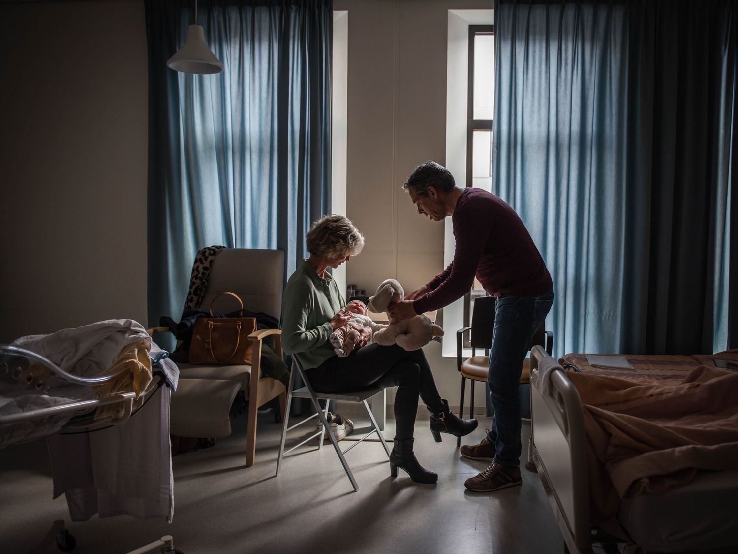 A man and woman sit in a hospital room, tenderly holding a newborn baby near a window with blue curtains.